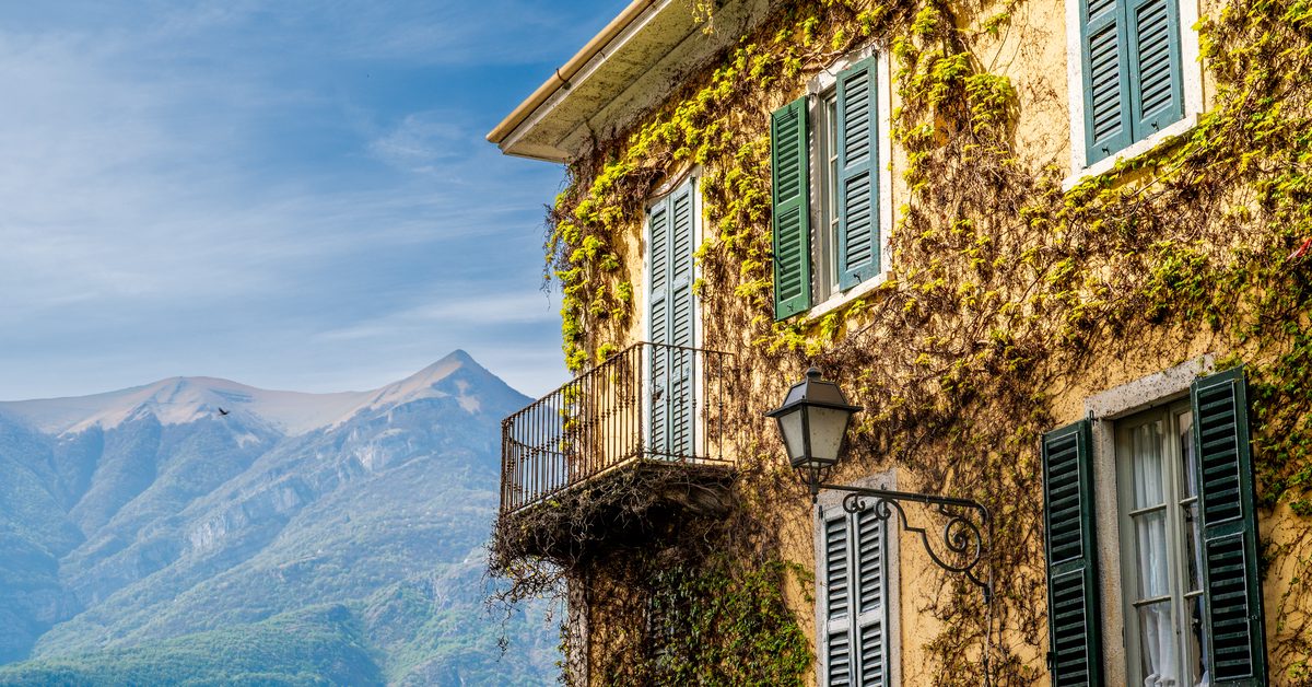 An ivy-covered villa with traditional green shutters overlooks the mountainous landscape surrounding Lake Como. Warm sunlight, weathered walls and Mediterranean vegetation give the scene a timeless northern Italian character.