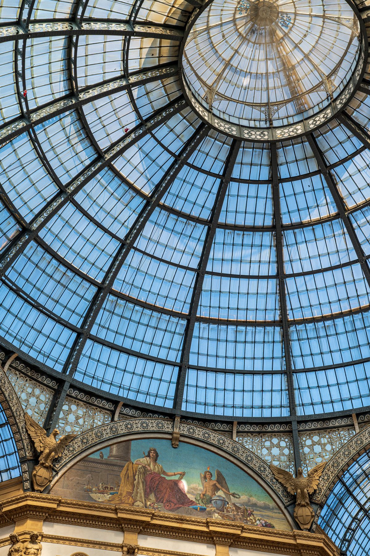 A view upward reveals the elegant iron-and-glass dome of Milan’s historic Galleria Vittorio Emanuele II. Ornamental metalwork, classical decoration and filtered spring sunlight highlight the grandeur of one of Italy’s most celebrated shopping arcades.