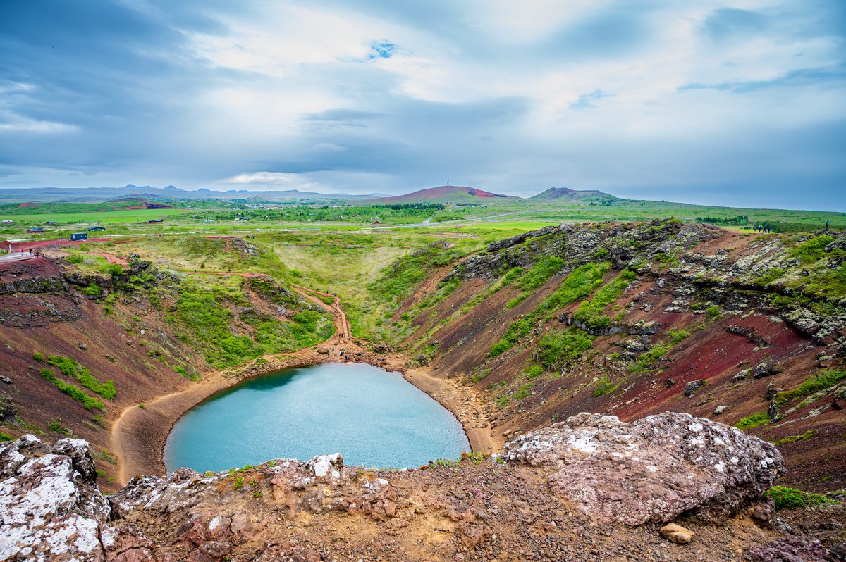 Standing on the jagged edge of the ancient crater, the view opens up past the striking blue waters to reveal a vast, rolling landscape stretching endlessly toward distant volcanic peaks.