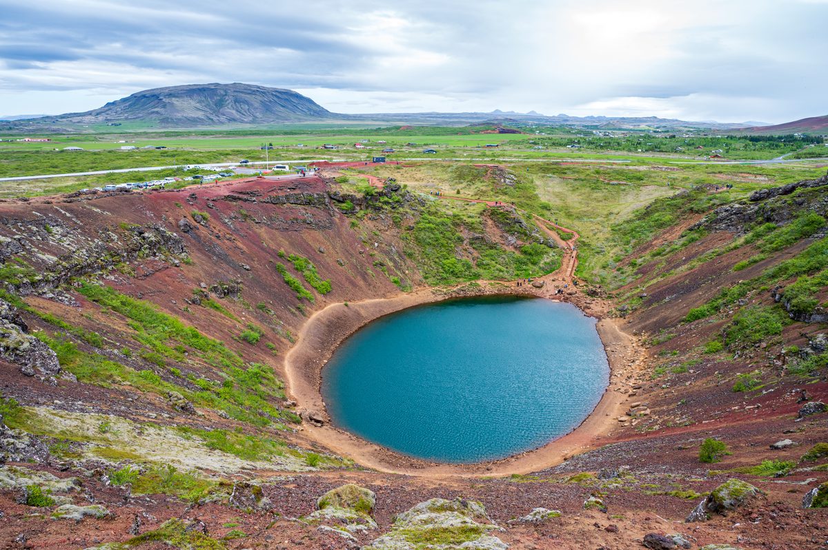 A high-angle panorama of a stunning volcanic crater lake, its vivid blue waters completely encircled by striking slopes of red earth, where tiny figures of travelers walk along the rim.