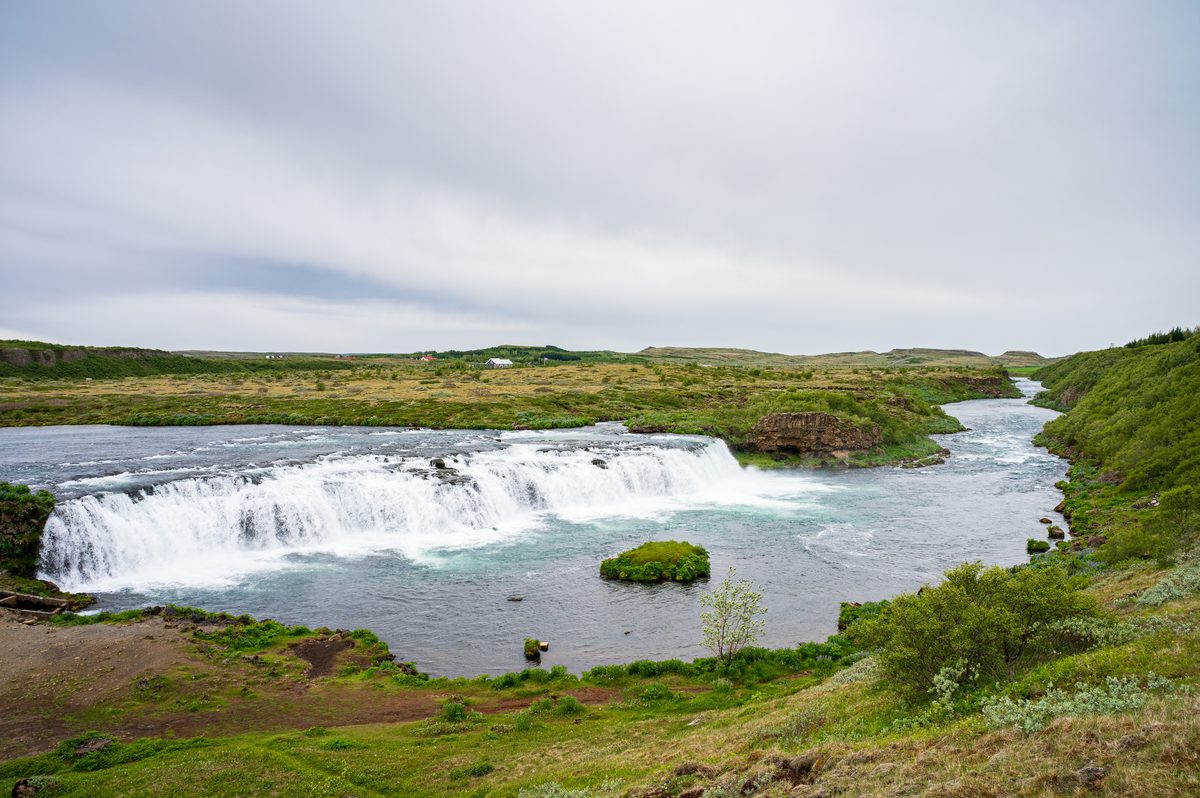 A breathtaking wide shot showing the river carving its way through the verdant valley before dropping over a broad, picturesque waterfall, highlighting the perfect harmony of land and flowing water.