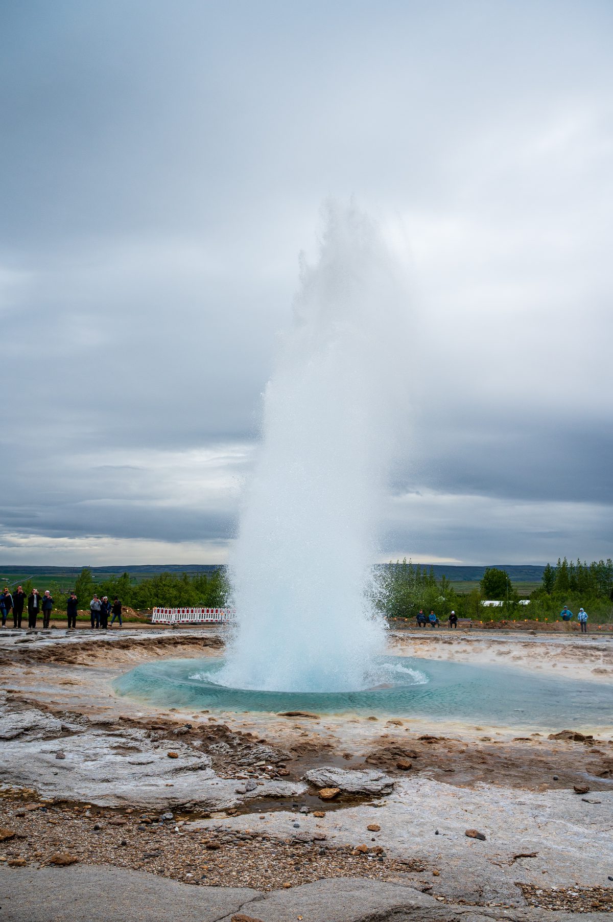A spectacular eruption of a geyser, launching a massive column of boiling blue water and white steam high into the cool air while onlookers watch nature's theatrical display.
