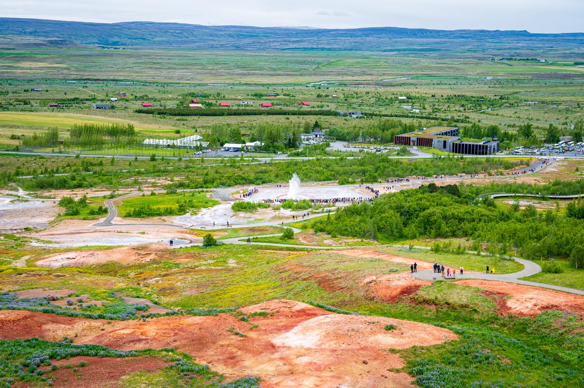 A high-angle view over a sprawling geothermal valley, capturing a geyser erupting as visitors gather along winding paths amidst vibrant red earth and green fields.