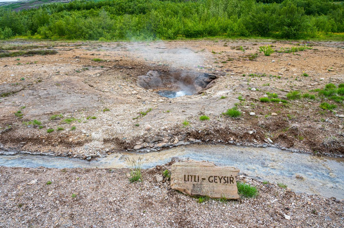 Steam rises gently from the bubbling waters of Litli-Geysir, a small but active geothermal spring set beside a shallow stream and rocky, mineral-stained earth.