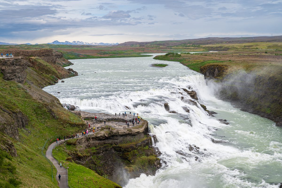 Tiny figures of travelers line the rocky viewing platform, offering a stunning sense of scale against the immense, roaring power of the wide river plunging into the jagged canyon.