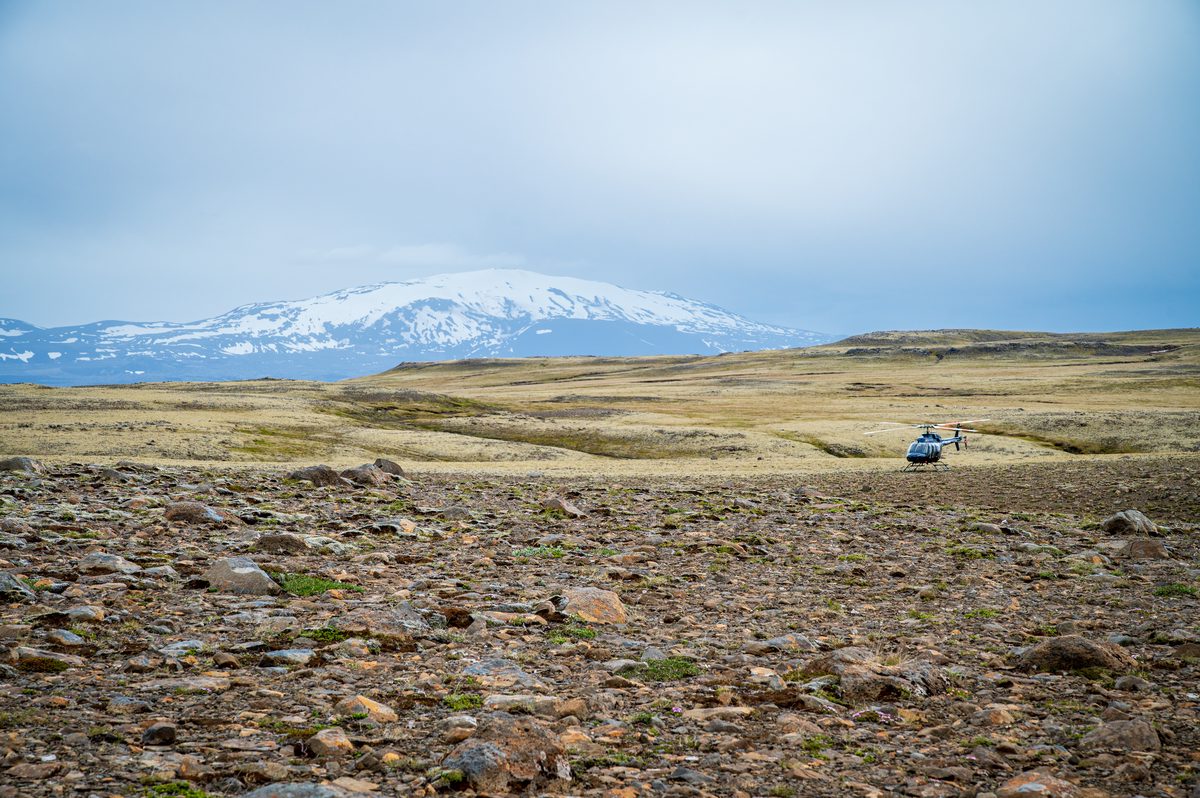 A lone helicopter rests on a rocky, desolate highland plain, completely dwarfed by the imposing, snow-capped presence of a massive volcanic peak rising in the distance.