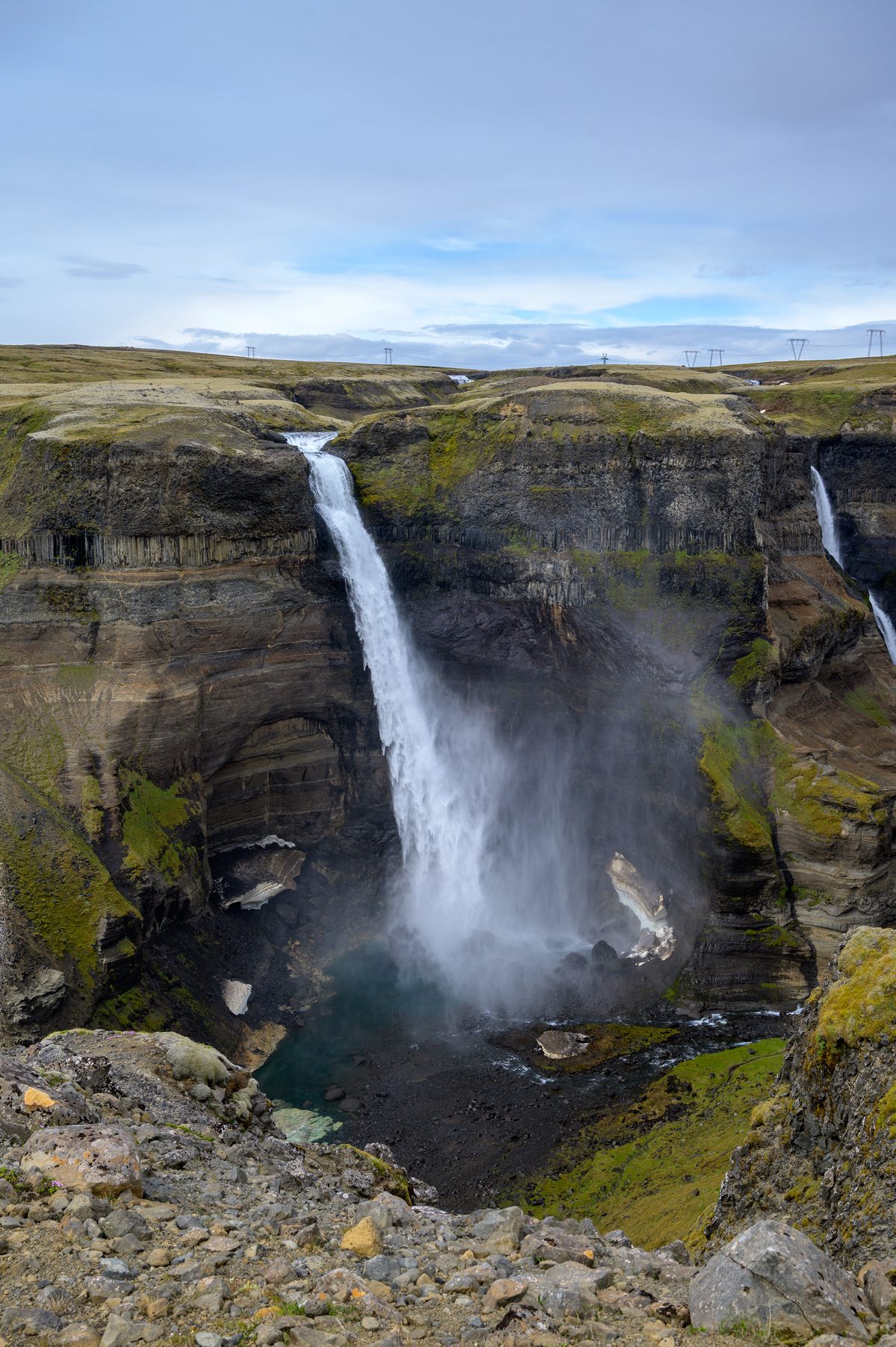 A dramatic, high-angle view looking straight down into the plunging basin of the great waterfall, where churning turquoise waters meet lingering patches of winter snow in the shadowy depths