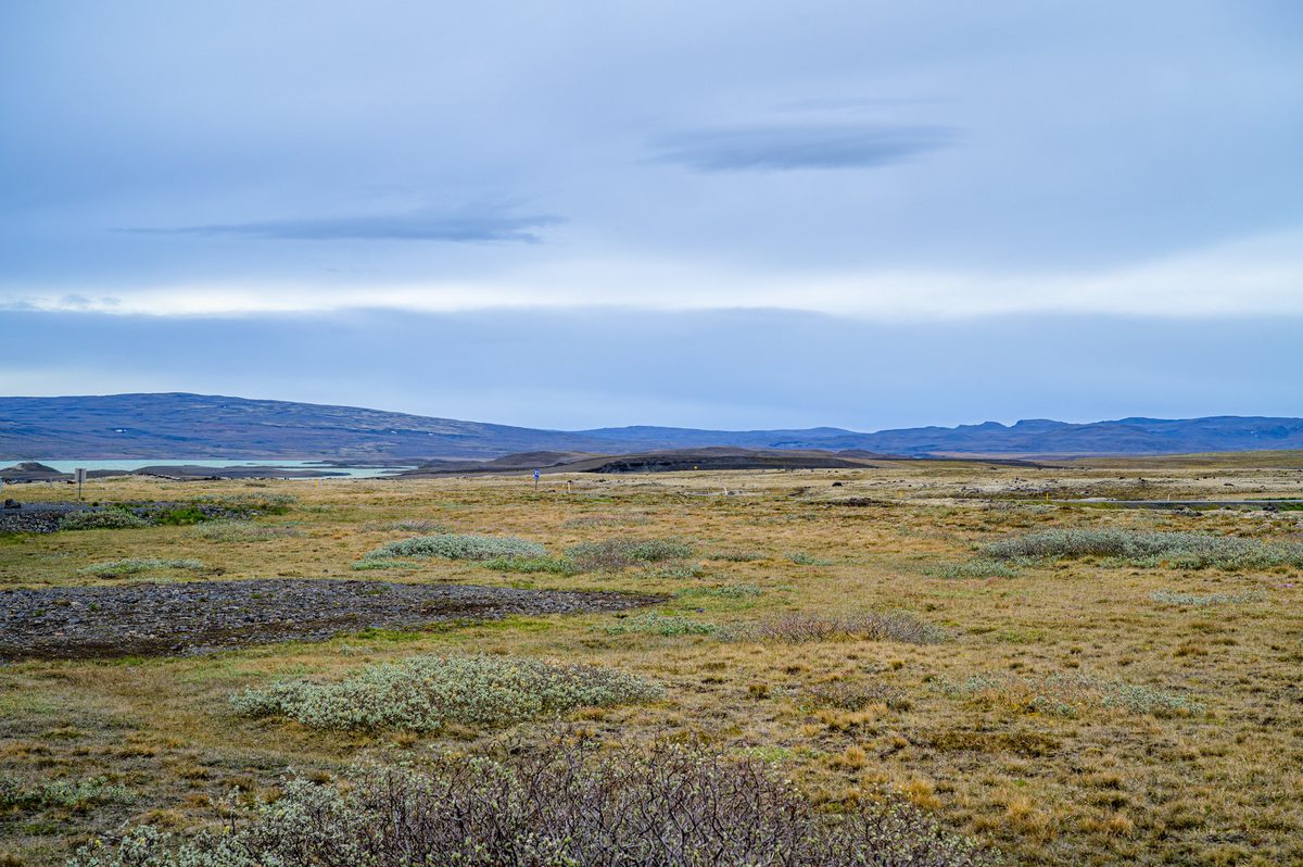 The desolate, sci-fi expanse of the highland plains stretches endlessly toward the horizon, where sparse brush, rocky earth, and a pale glacial lake define the stark, beautiful isolation of the route to Sprengisandur.