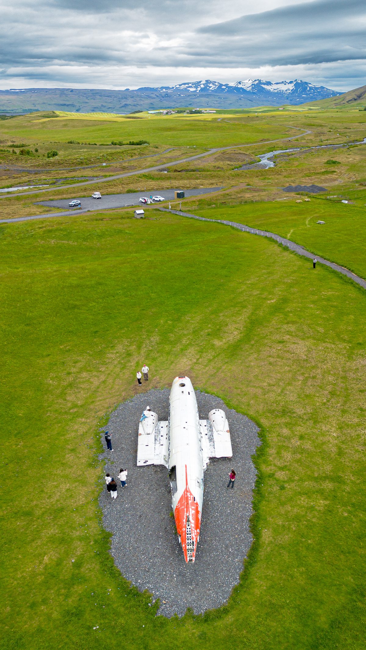 An aerial view of the abandoned aircraft wreck at Sólheimasandur, surrounded by vivid grass, winding tracks and distant snow-streaked mountains.