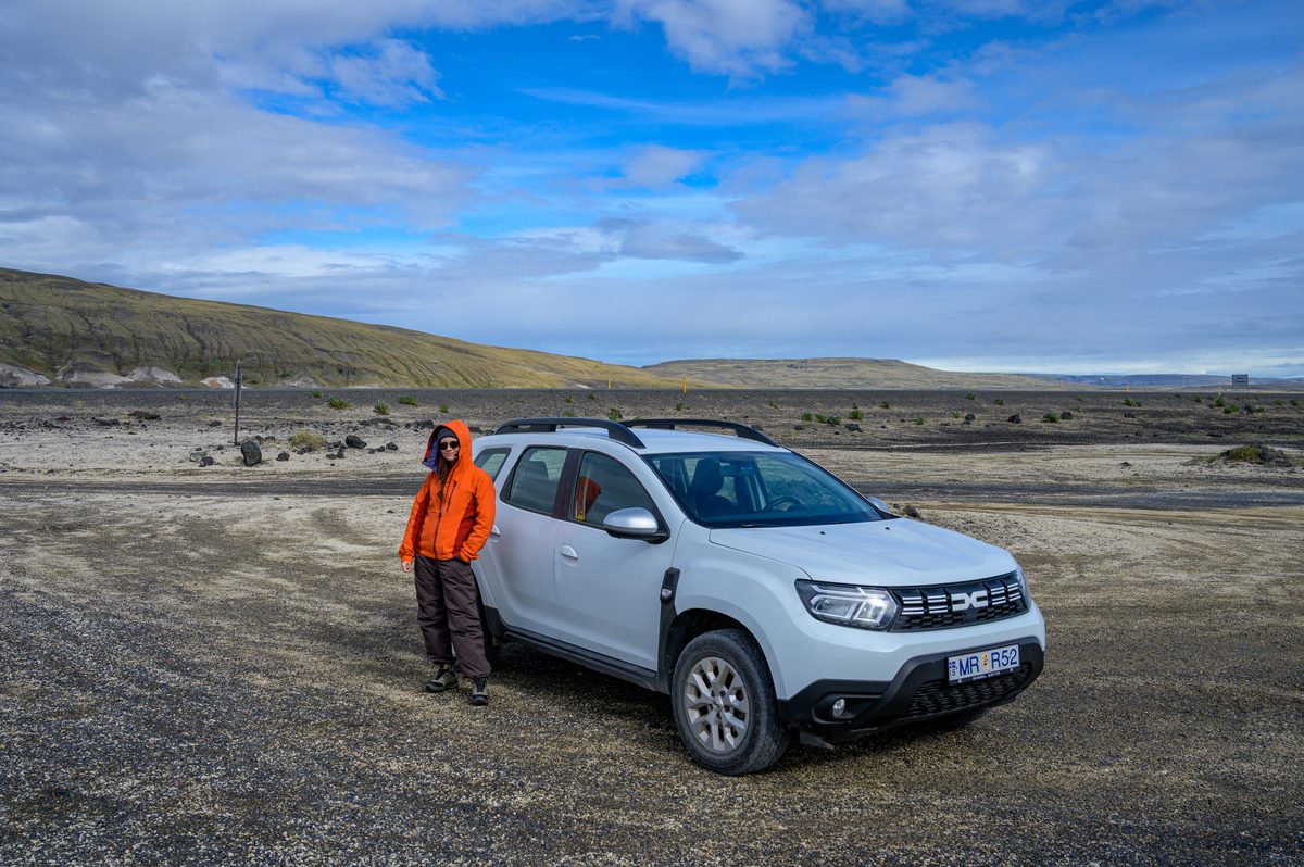 A traveler in a bright orange jacket stands beside the day’s white rental car on a stark volcanic plain beneath a clearing blue sky.