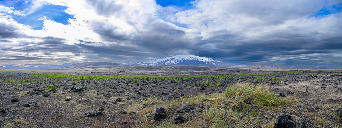A panoramic view stretches across a young green-speckled lava field toward snow-covered Hekla beneath sweeping layers of light and storm cloud.