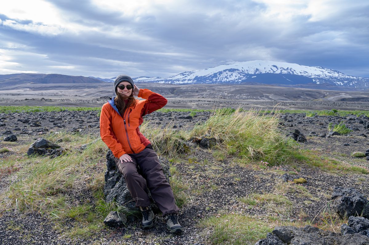 Wrapped against the wind in an orange jacket, a traveler smiles from a lava rock with snowy Hekla rising across the open plain.