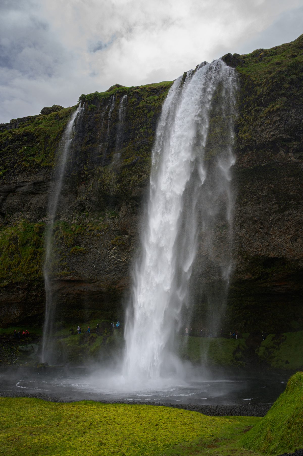 Seljalandsfoss plunges from a moss-covered cliff into a basin of spray, with tiny visitors tracing the path behind the water.