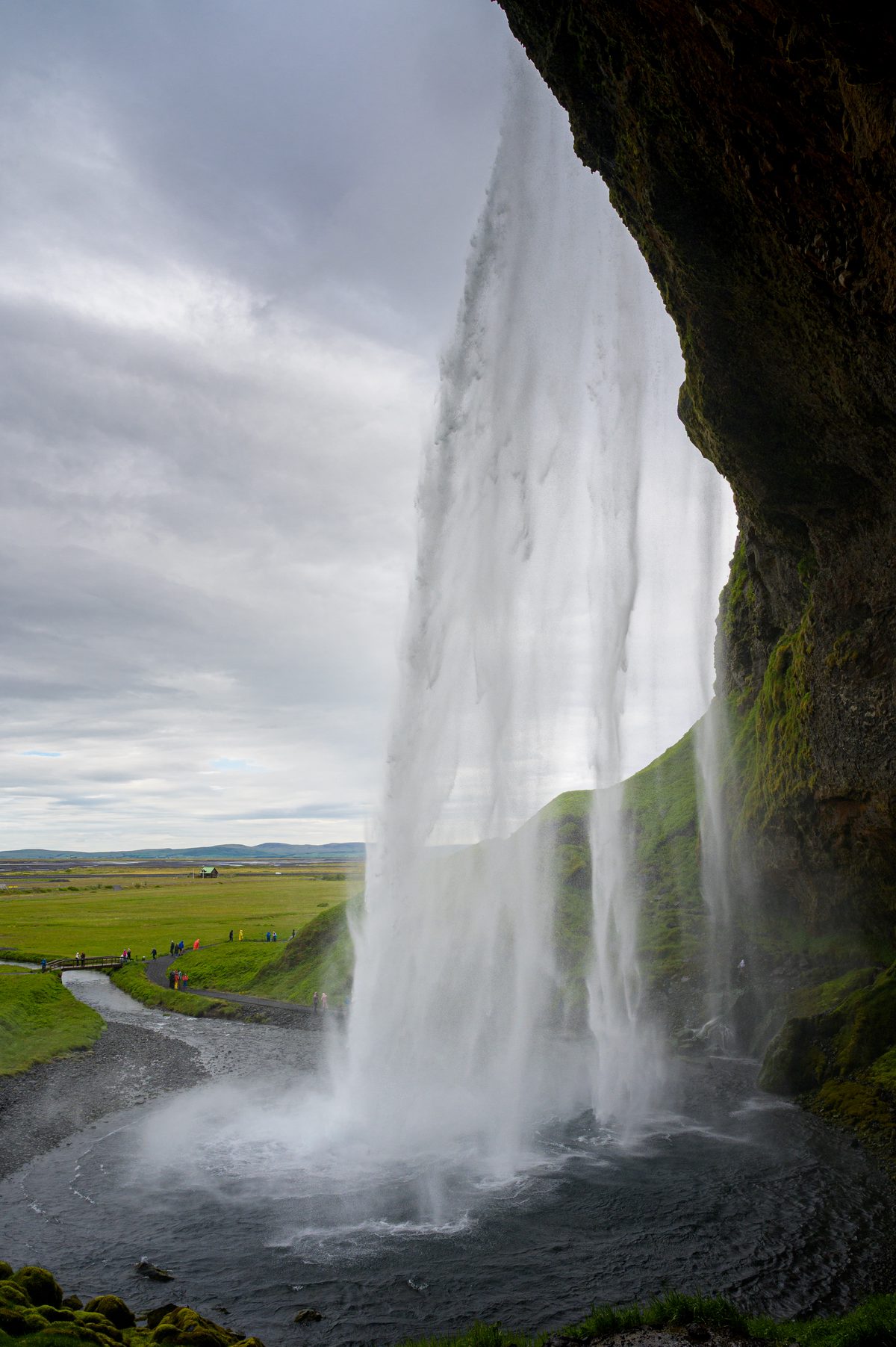 From the cavern behind Seljalandsfoss, the waterfall forms a translucent curtain over the dark pool and green lowlands beyond.