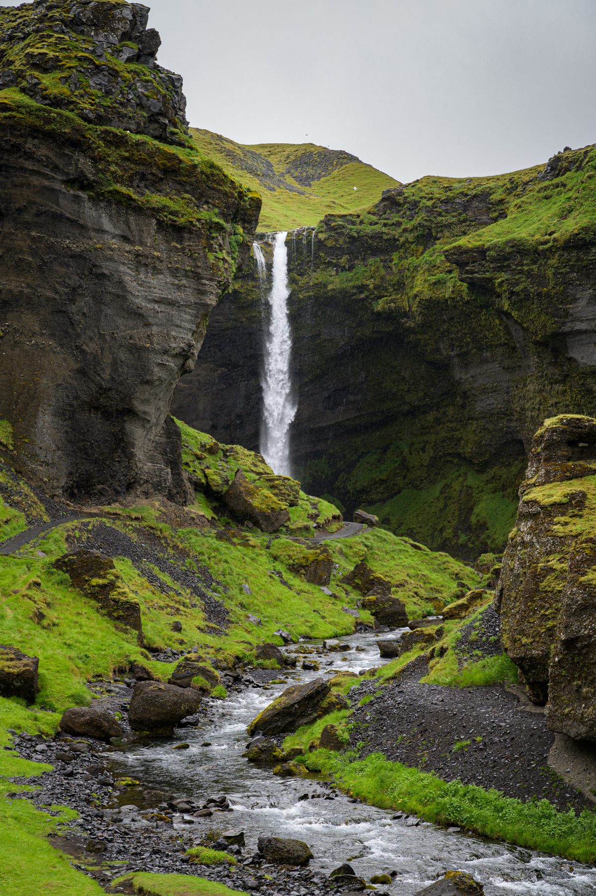 Kvernufoss drops into a secluded moss-green ravine as a rocky stream winds toward the foreground.
