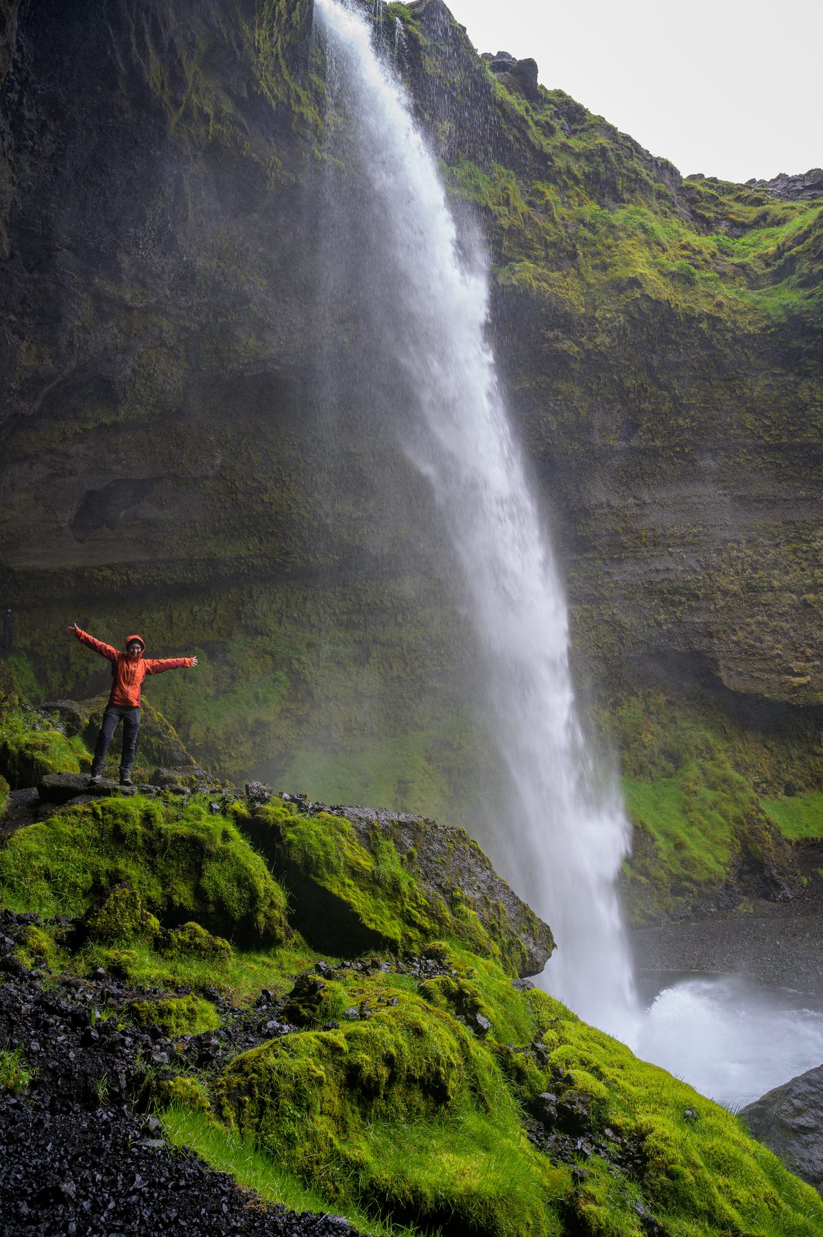 A traveler in orange raises her arms from a mossy ledge beside the tall ribbon of Kvernufoss.