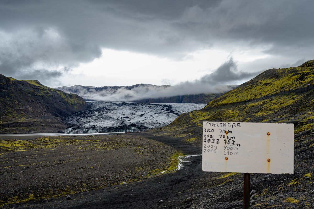 A hand-painted sign records the changing distance to Sólheimajökull, whose ash-streaked ice emerges between dark slopes and low cloud.