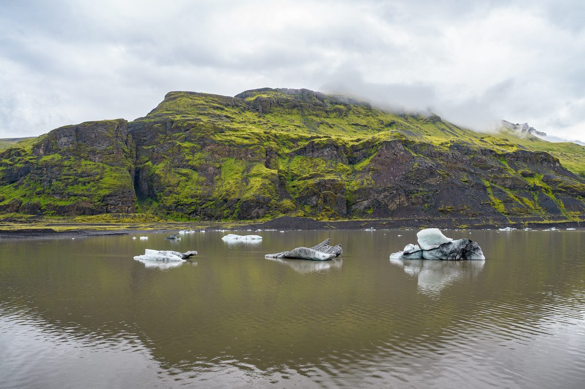 Small blue-white icebergs drift across the still brown water of the Sólheimajökull glacial lagoon below luminous green slopes.