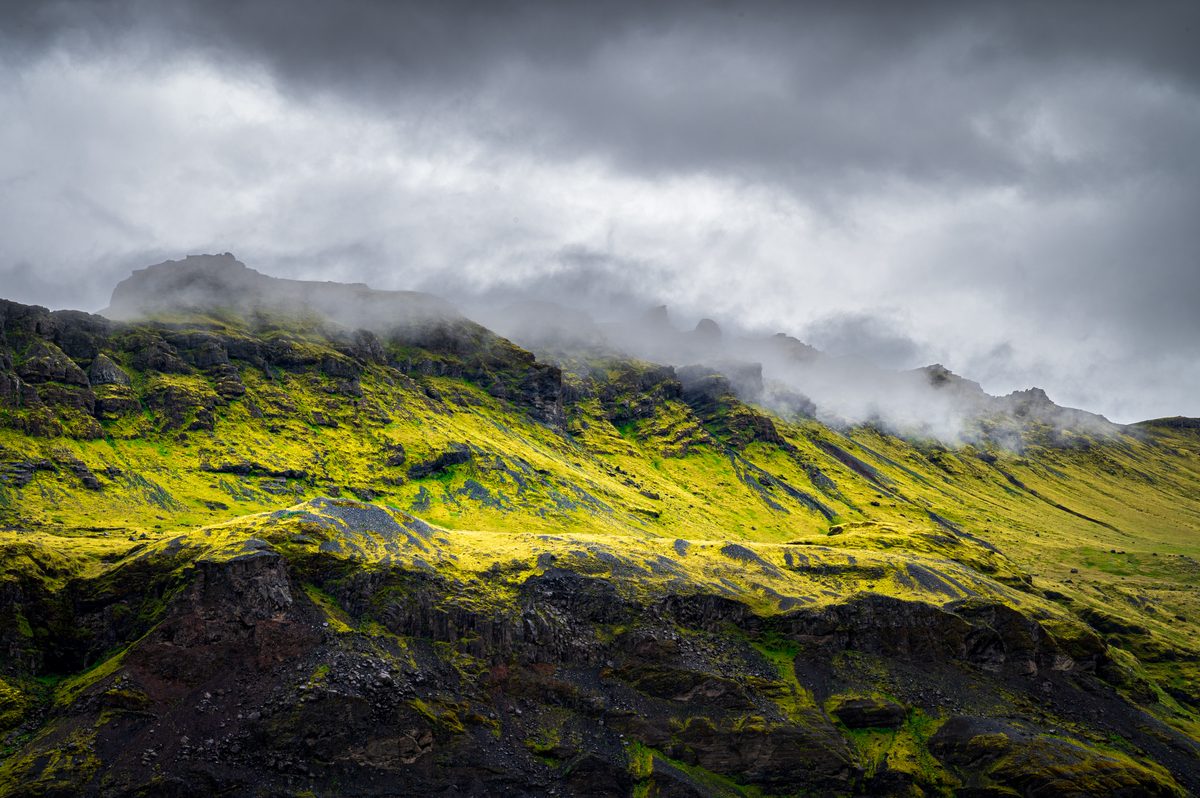 A shaft of light ignites the green slopes above Sólheimajökull while mist curls beneath a dark, restless sky.