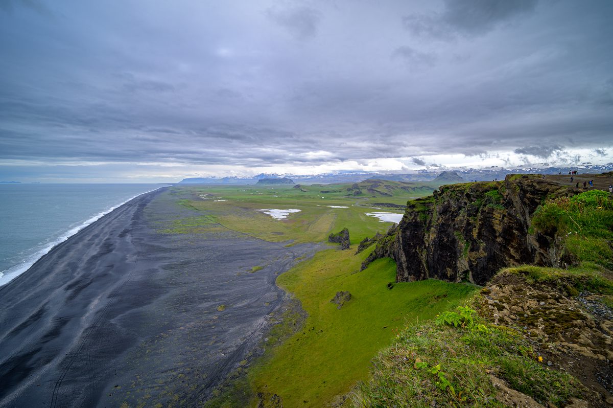From Dyrhólaey’s cliffs, a sweeping black-sand shoreline runs toward distant snow-covered mountains beneath a stormy sky.