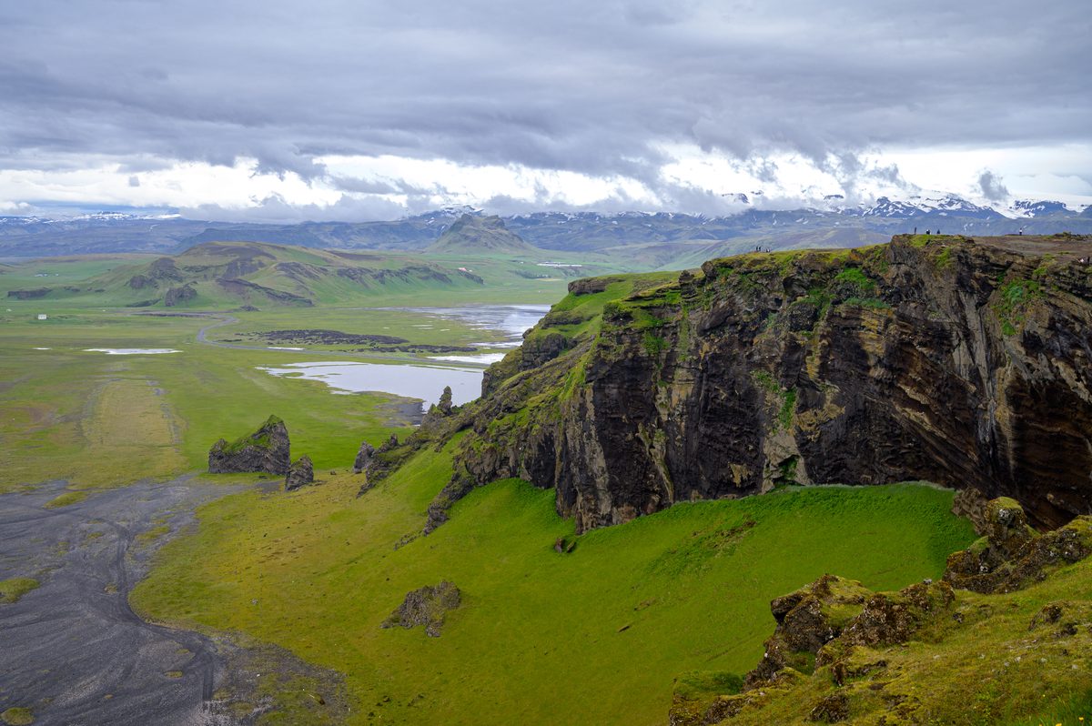 Jagged green cliffs fall toward wetlands and black sand, with distant mountains gathered beneath bands of cloud.