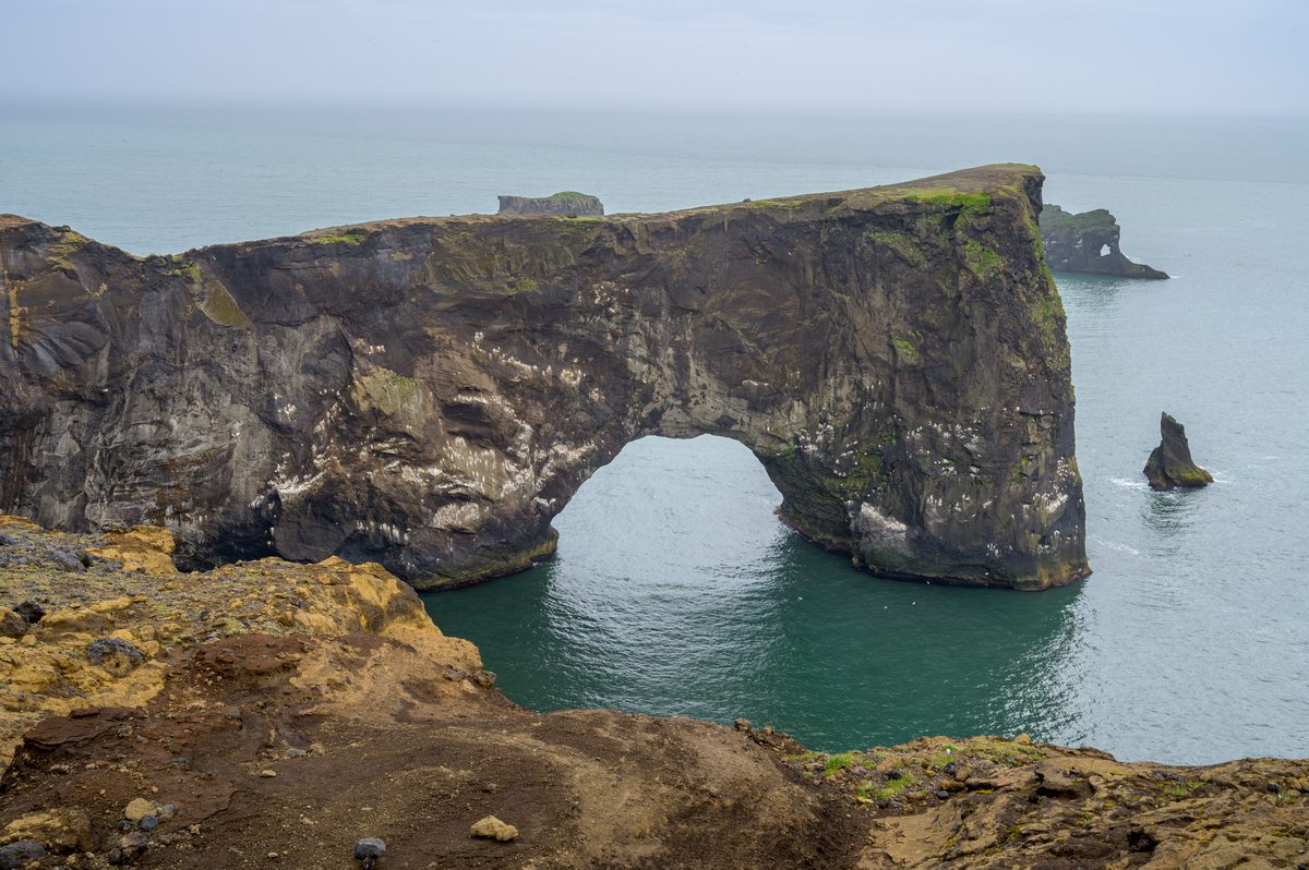 Dyrhólaey’s massive sea arch rises from turquoise water, with rock stacks and seabirds scattered around the weathered cliffs.