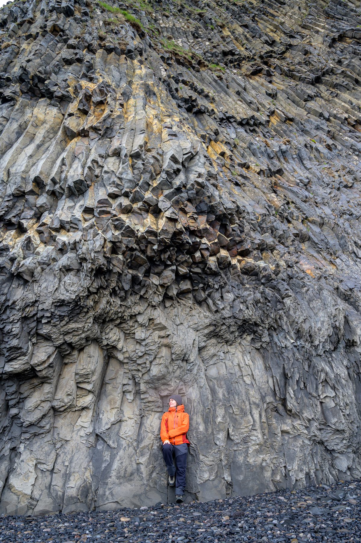 A traveler in orange looks up at the towering, twisted basalt columns along Iceland’s black-pebble coast.