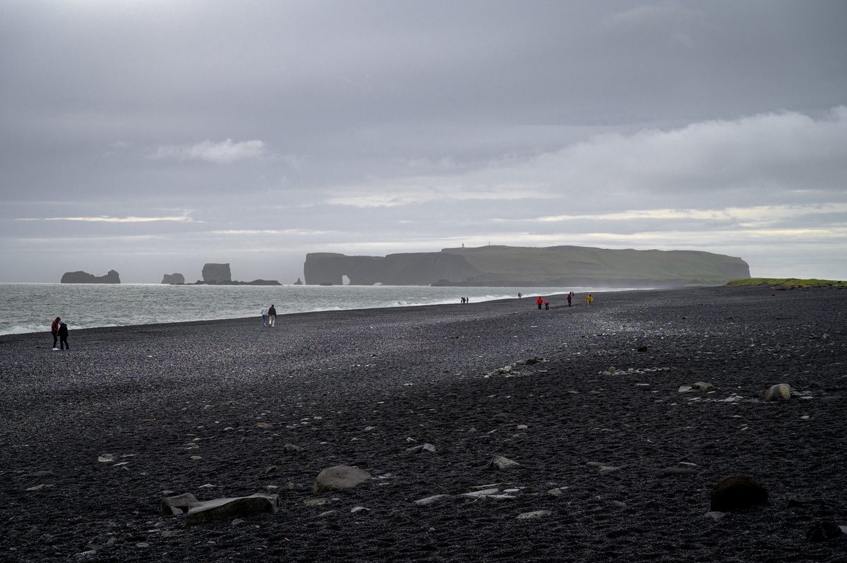 Visitors cross a broad black-pebble shore while the distant Dyrhólaey headland and its sea arch fade into coastal rain.