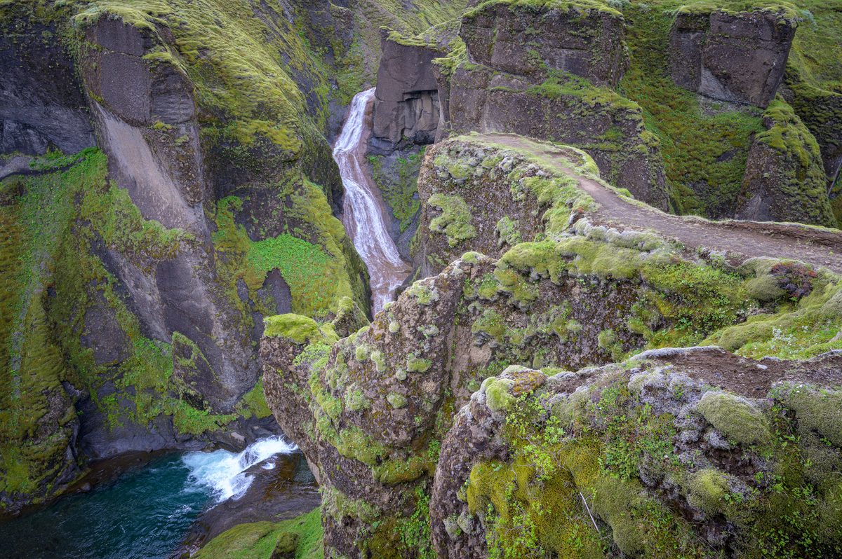 A narrow moss-lined path follows the sculpted edge of Fjaðrárgljúfur above a waterfall and turquoise pool.