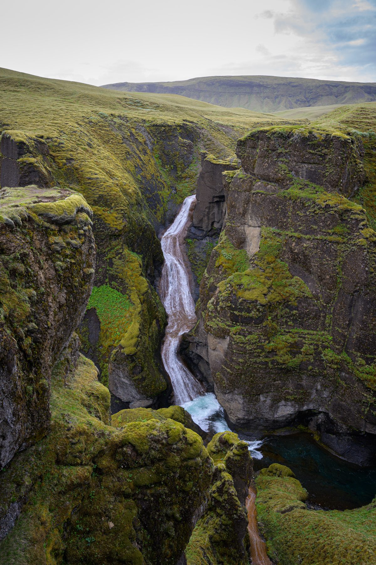 A twisting waterfall descends through the tall green-and-black walls of Fjaðrárgljúfur into a deep blue pool.