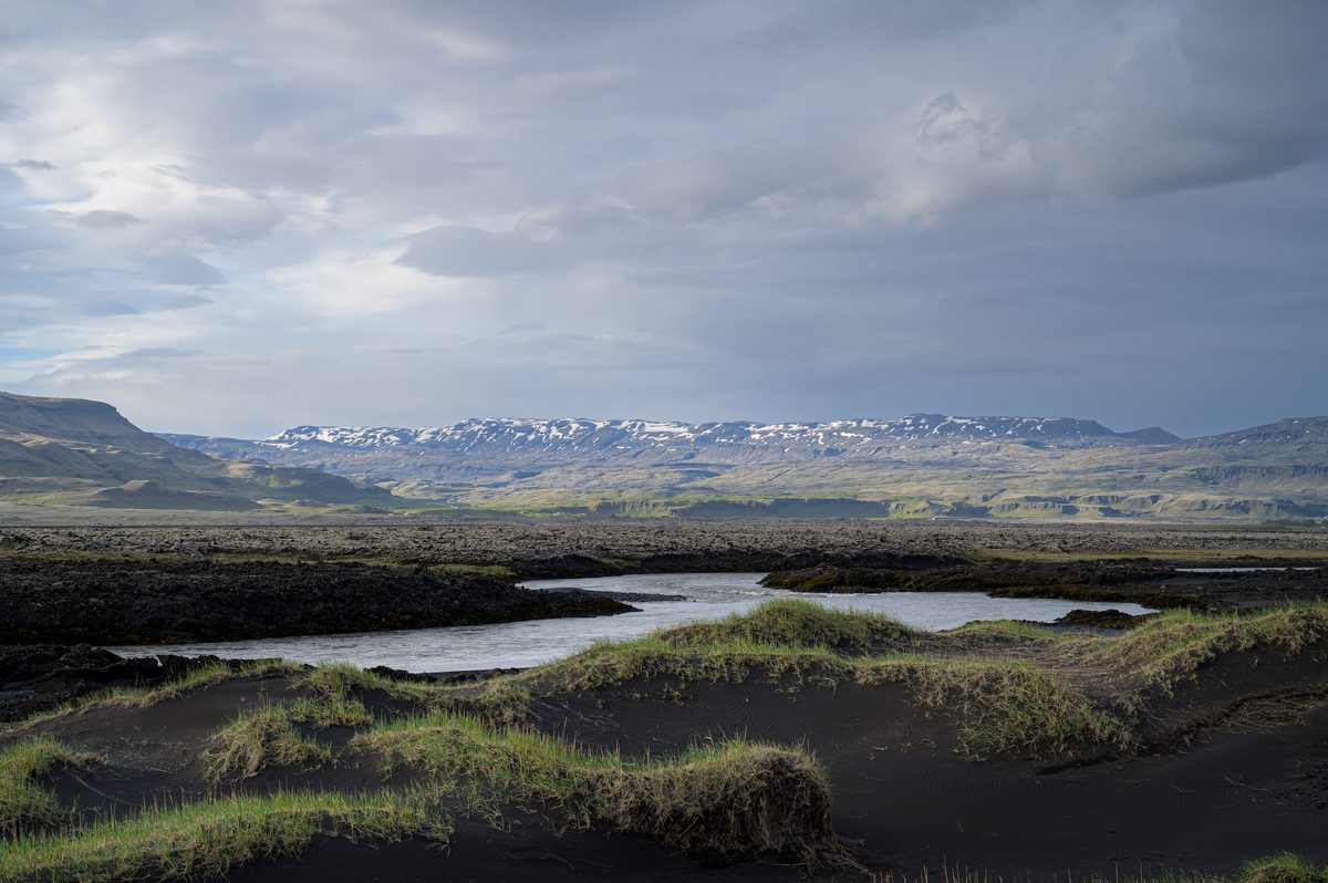 Wind-shaped black-sand hummocks frame a quiet braided pool, with snow-patched ridges glowing beneath broken cloud.