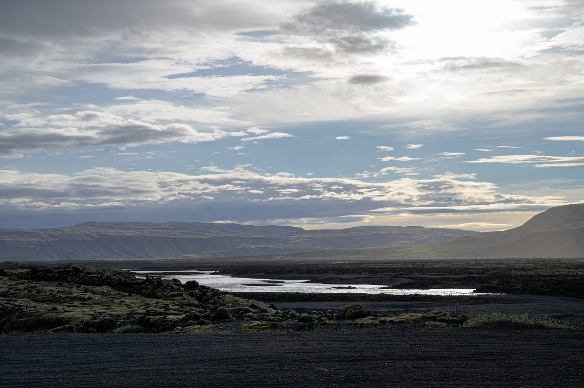 Late light catches the bends of a river crossing the dark volcanic plain beneath a luminous, cloud-filled sky.