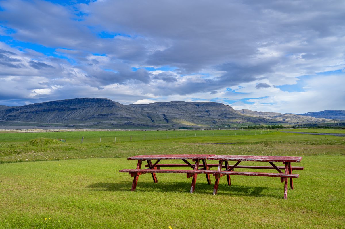 Red picnic tables sit in a vivid green field beneath layered mountains and a shifting Icelandic sky.