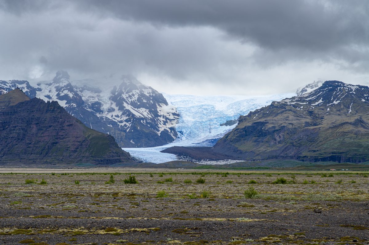 A pale glacier tongue flows between dark, snow-streaked mountains beneath low storm clouds.
