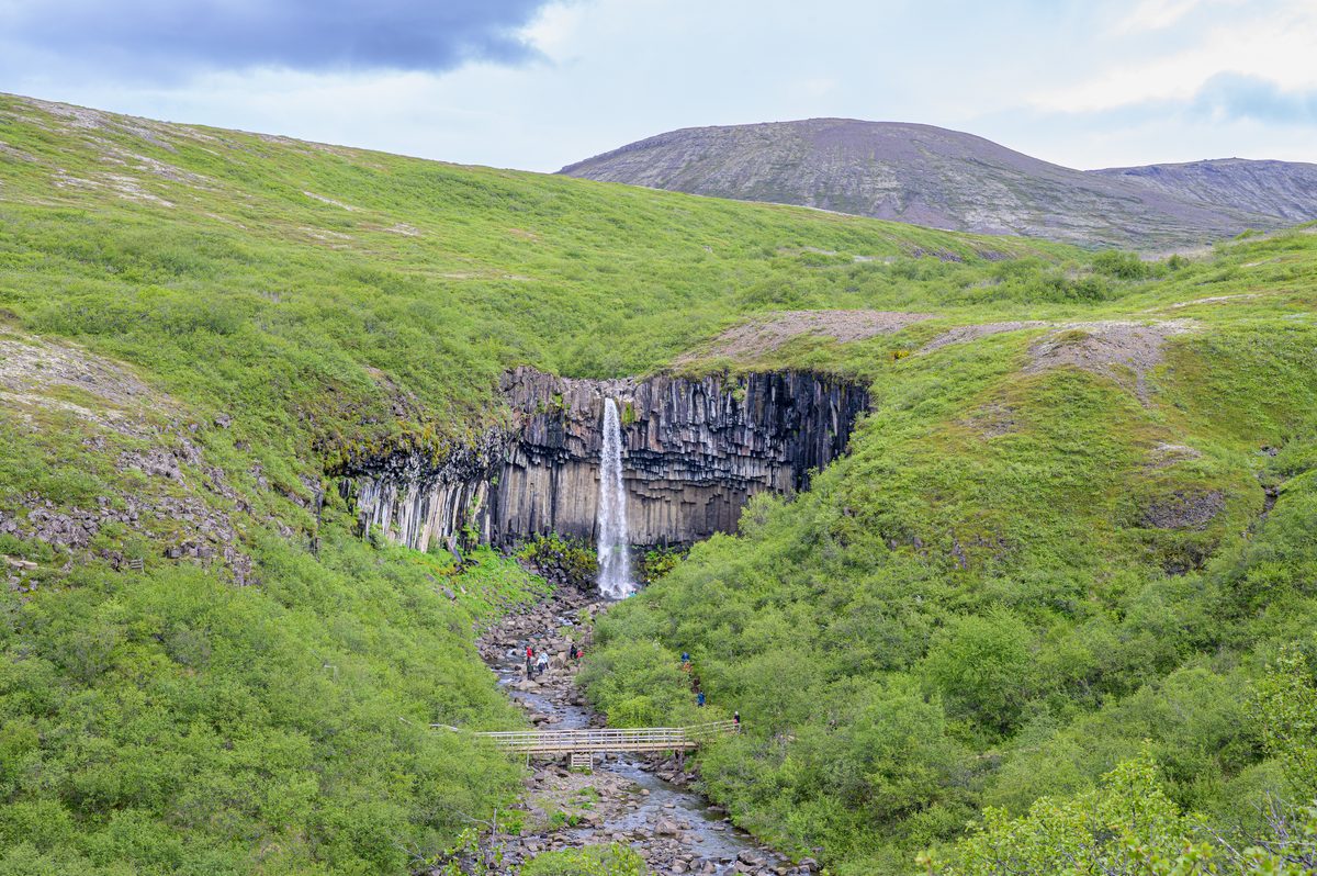 Svartifoss appears at the end of a green valley, framed by dark basalt and rolling hills.