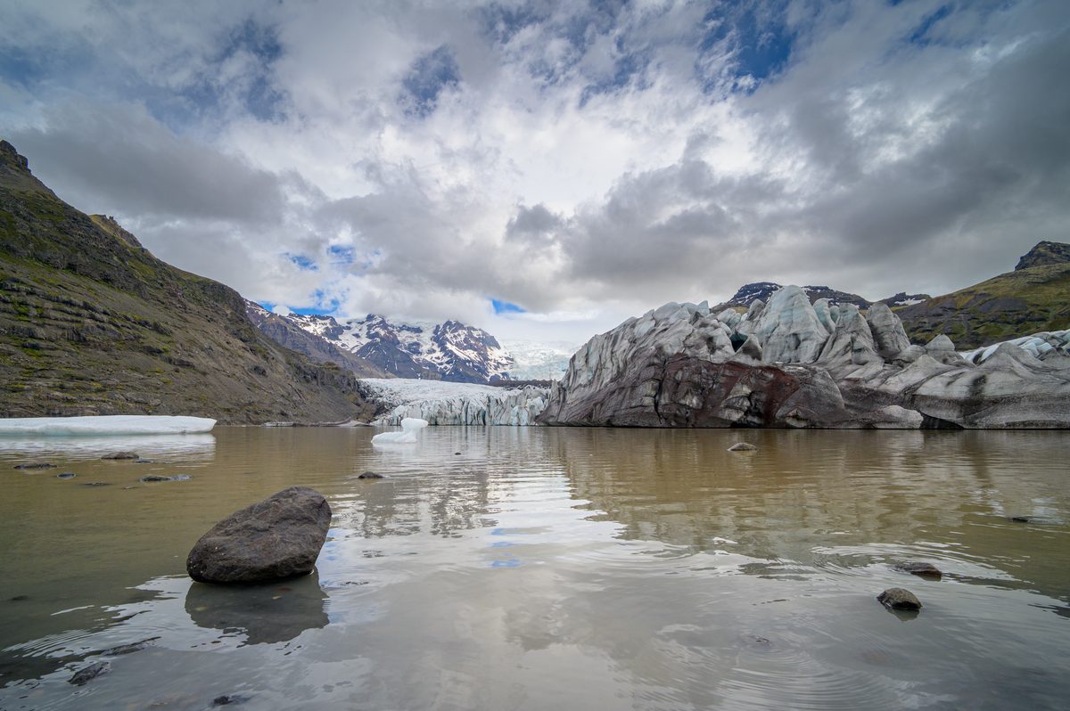 Weathered glacier ice and rounded stones are reflected in the calm, silty water of a secluded lagoon.