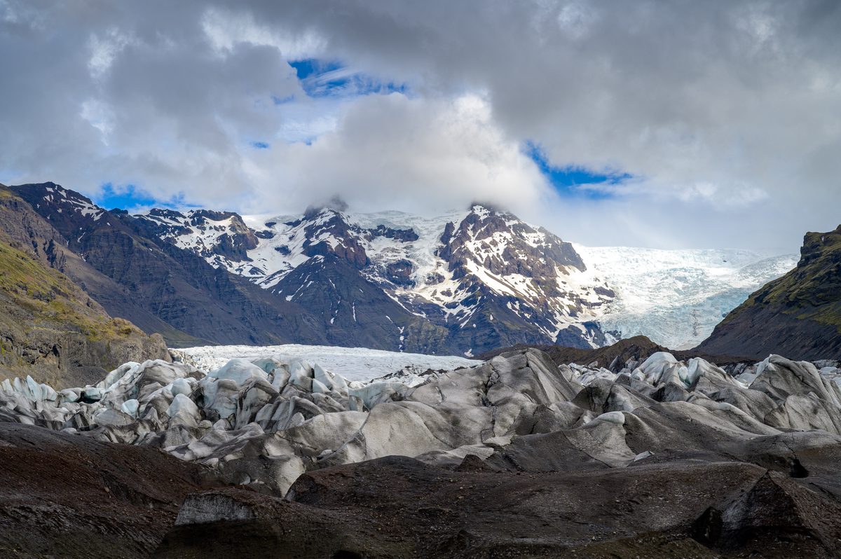 Jagged, ash-striped ice fills the foreground below snow-streaked peaks and a bright window in the clouds.