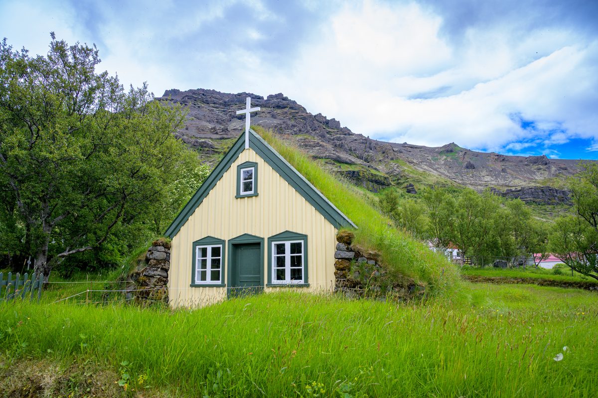 The turf-roofed Hofskirkja nestles into tall grass beneath a rocky ridge and luminous summer sky.