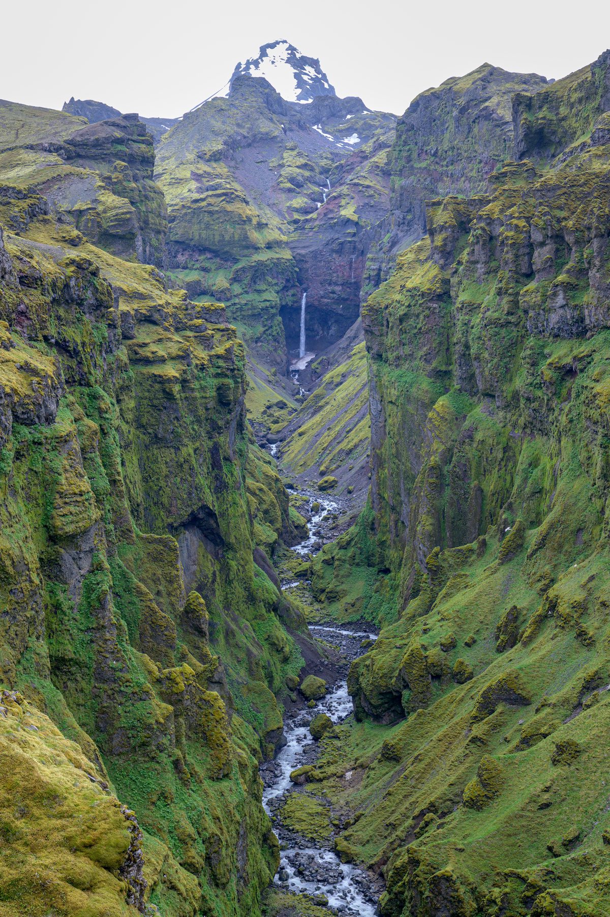 A distant waterfall and rushing stream draw the eye deep into the canyon’s shadowed, moss-covered walls.