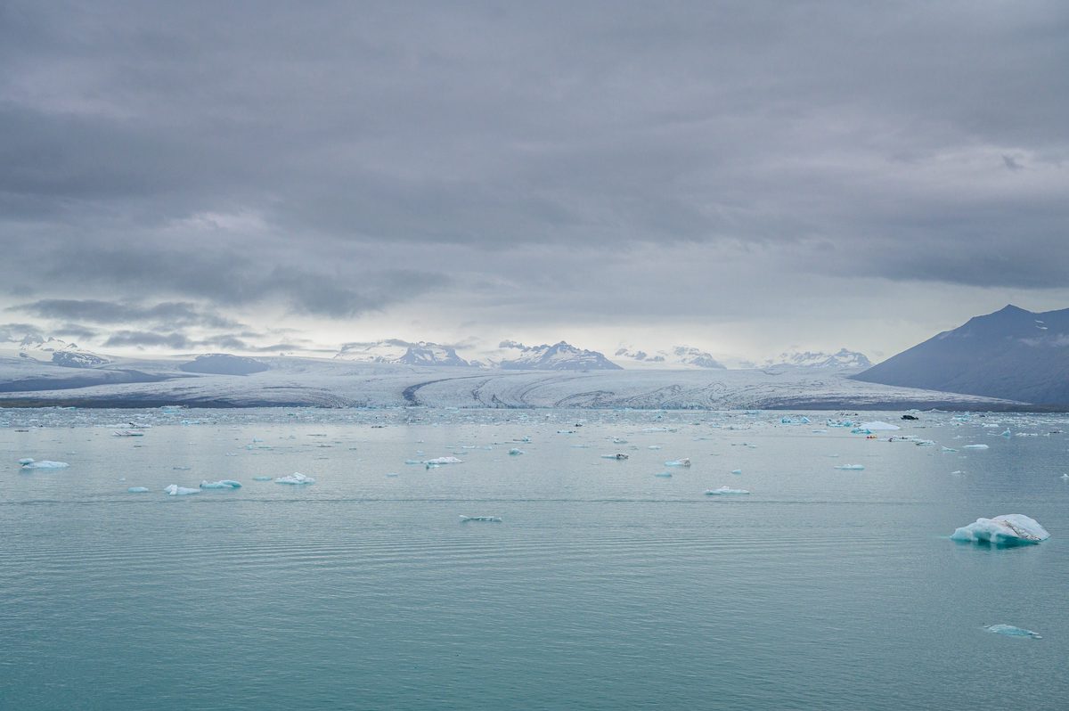 Small icebergs scatter across the quiet lagoon below distant glaciers and cloud-wrapped mountains.