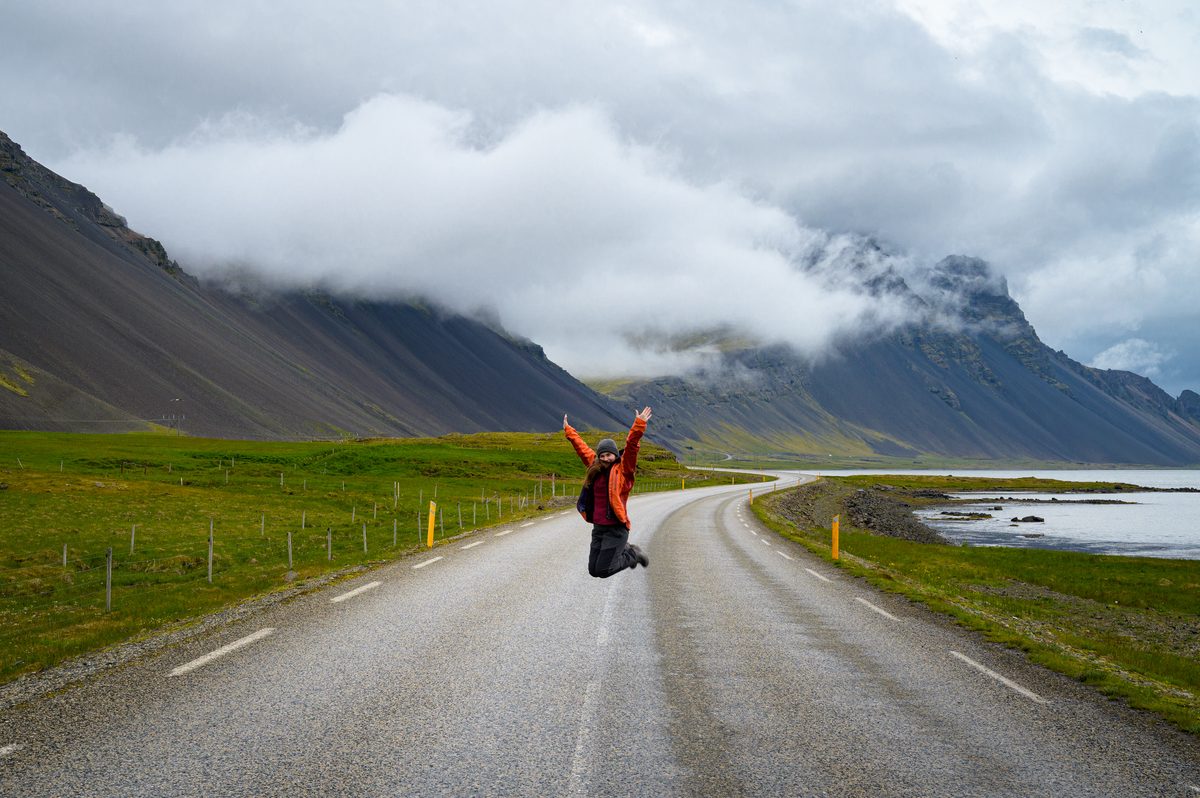 A traveler leaps in celebration on an empty road winding between a quiet inlet and cloud-covered mountains.