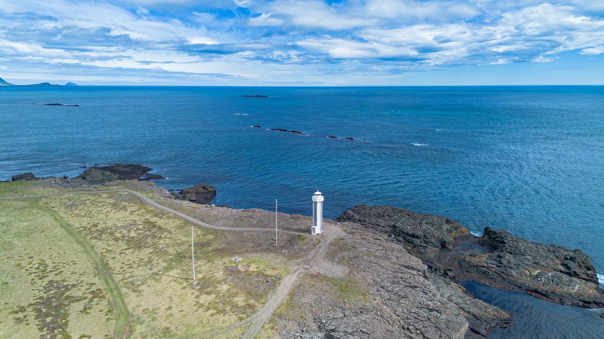 A white lighthouse stands on a dark rocky point where the calm blue sea opens beneath a sweeping summer sky.