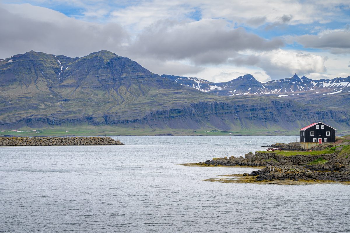 A solitary black house with white trim sits at the water's edge beneath the immense layered mountains of Berufjörður.
