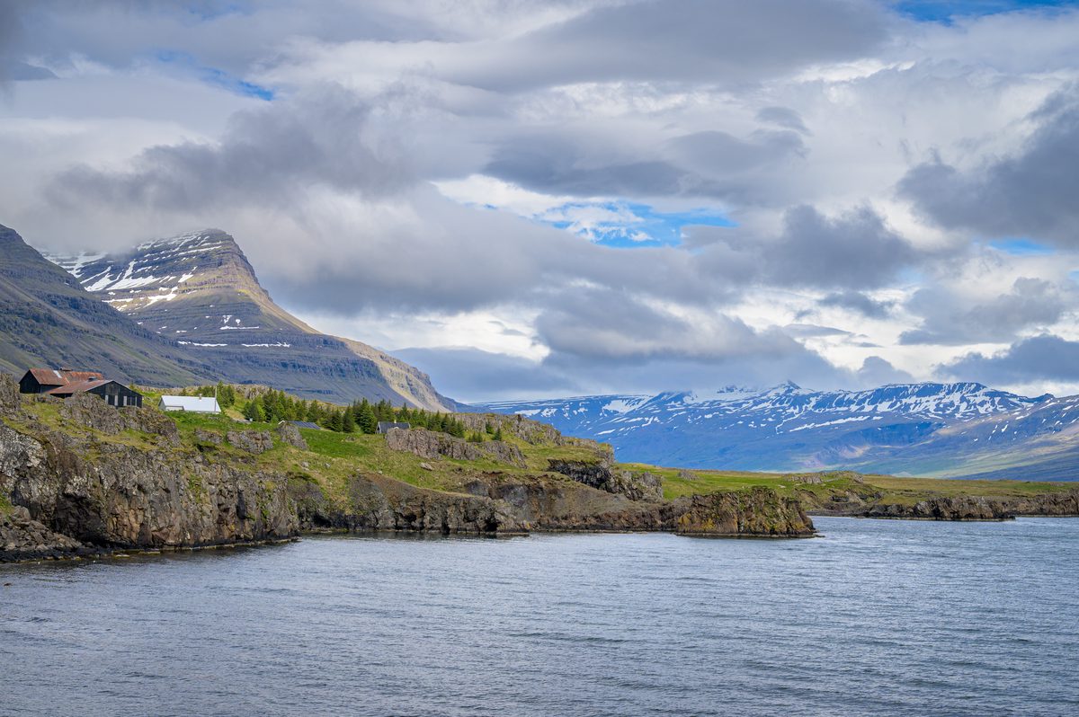 Small farm buildings cling to a rocky green shore while snow-striped ridges recede beneath shifting cloud.