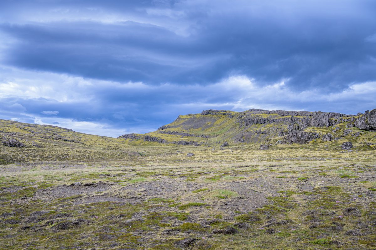 A sparse highland meadow rises toward fractured basalt ledges beneath heavy blue-gray storm clouds.