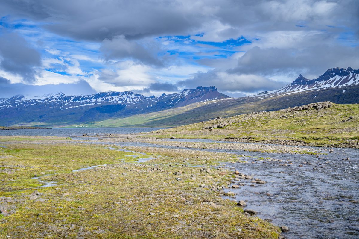 A clear stream crosses a stony valley toward the fjord and the jagged mountain wall beyond.