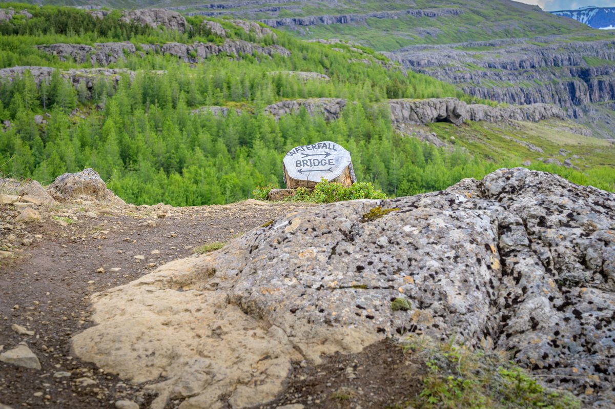 A hand-painted sign points toward the bridge and waterfall trail among rock, birch, and mountain terraces.
