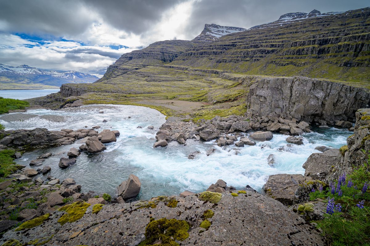 The milky-blue river curves around boulders beneath a steep terraced mountain before opening toward the fjord.
