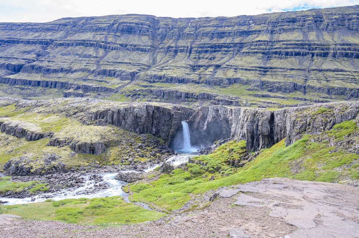 A broad waterfall drops into a green hollow surrounded by sheer dark cliffs and repeating volcanic terraces.