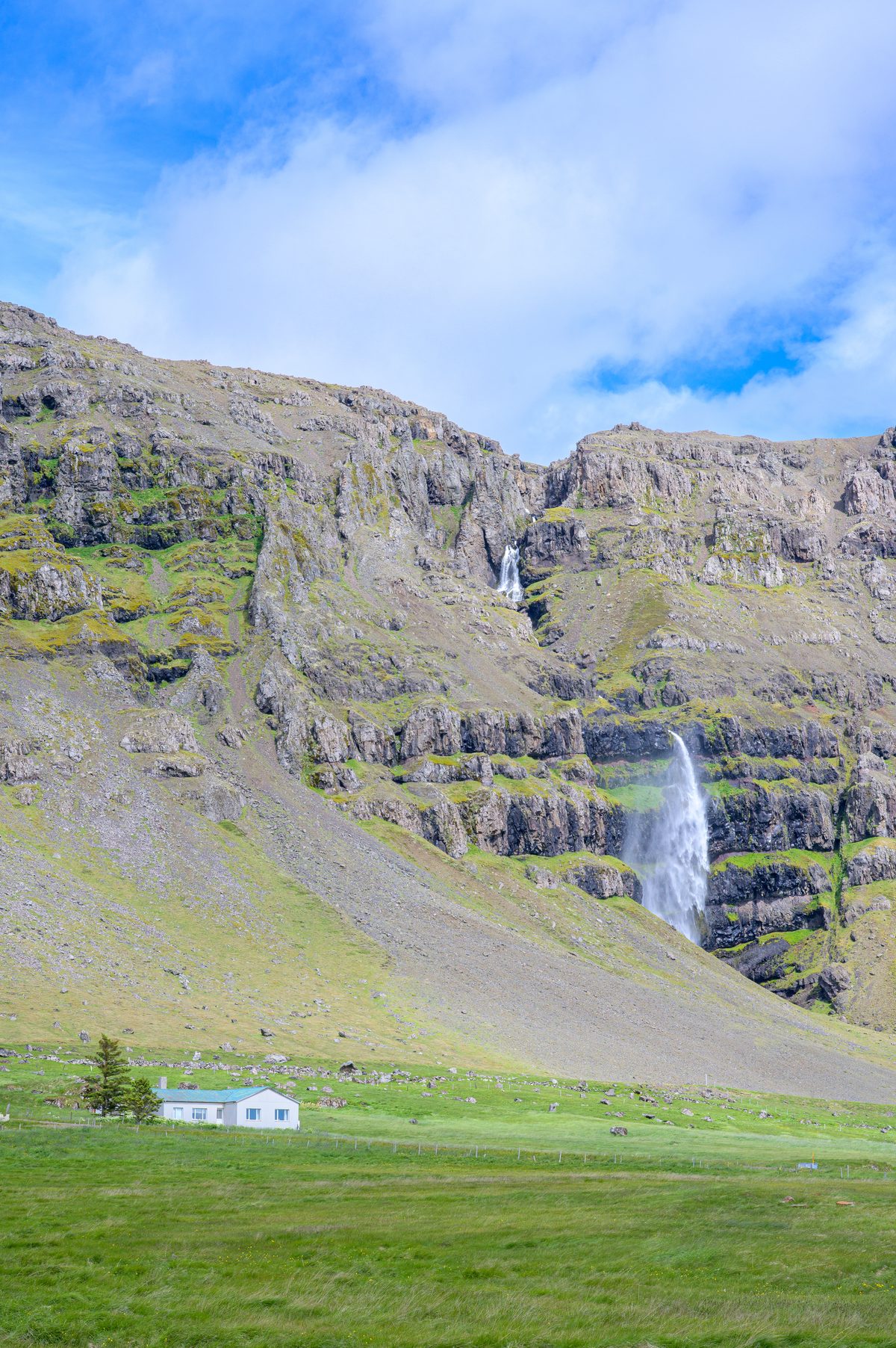 Two waterfalls descend a high mountain wall above a small white farmhouse and vivid green pasture.
